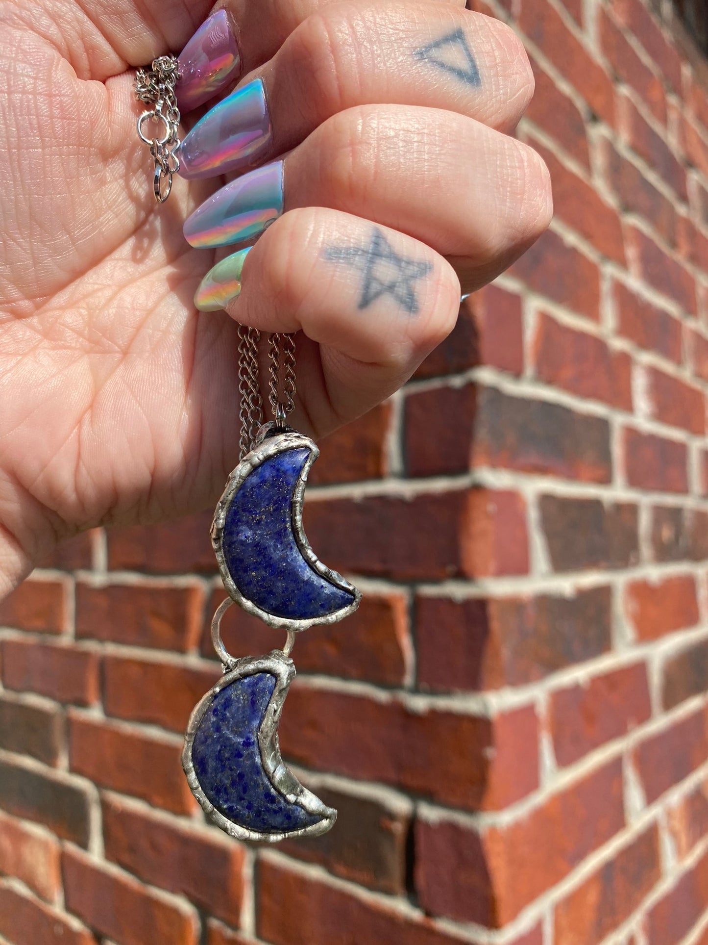 hand holding two lapis lazuli crescent moon pendants in front of a brick wall